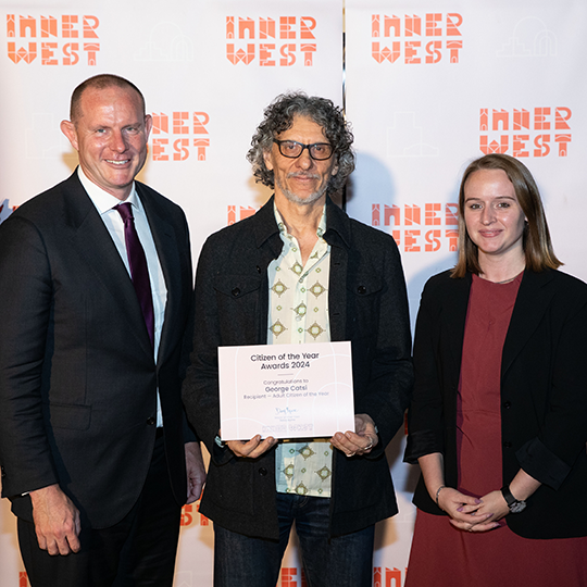 Three people in from of an Inner West COuncil meida wall. The person in the middle holds a certificate that reads: 'Citizen of the Year Award'